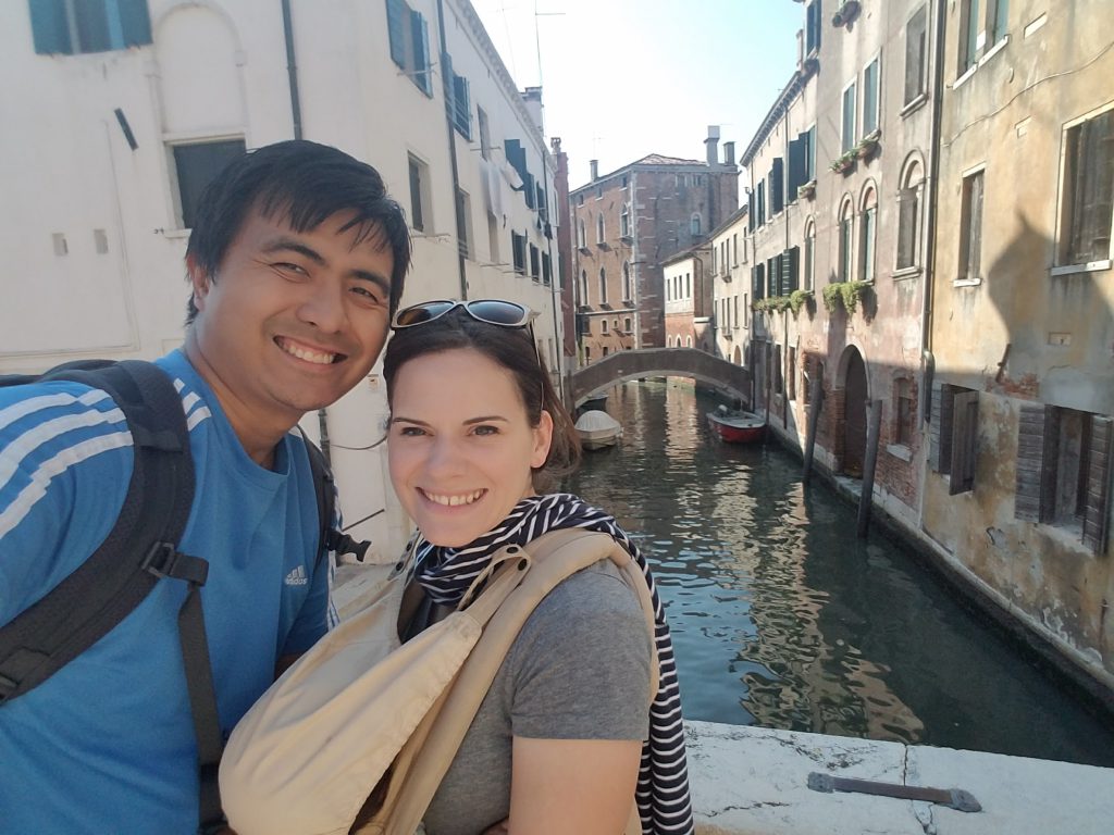 Man and woman in front of a canal in Venice, woman is wearing a sleeping baby in a baby carrier