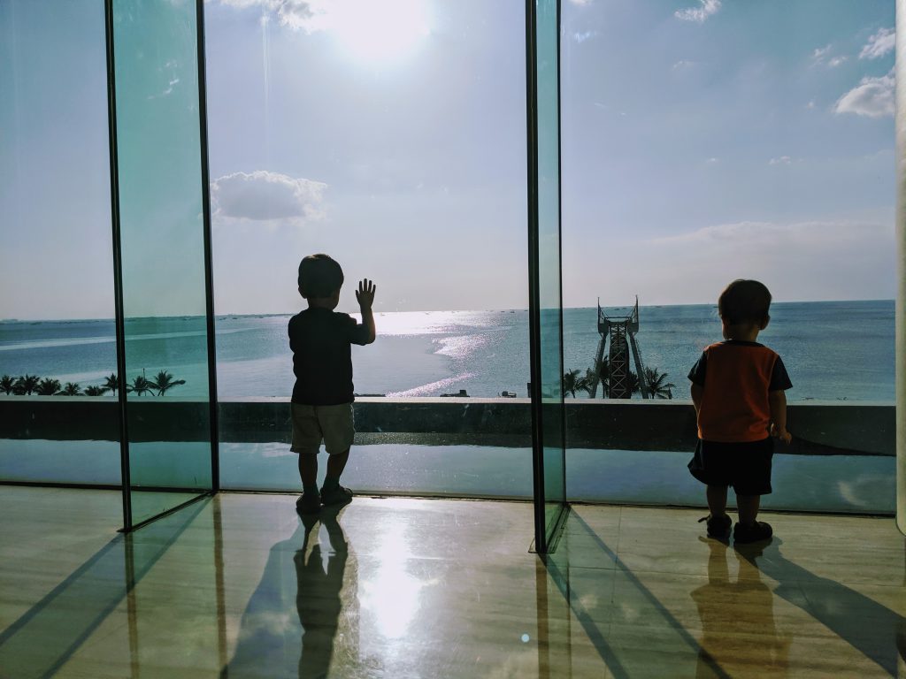 Two children looking out a window at a sunny sea.  Sunshine helps fight jet lag in babies and toddlers.